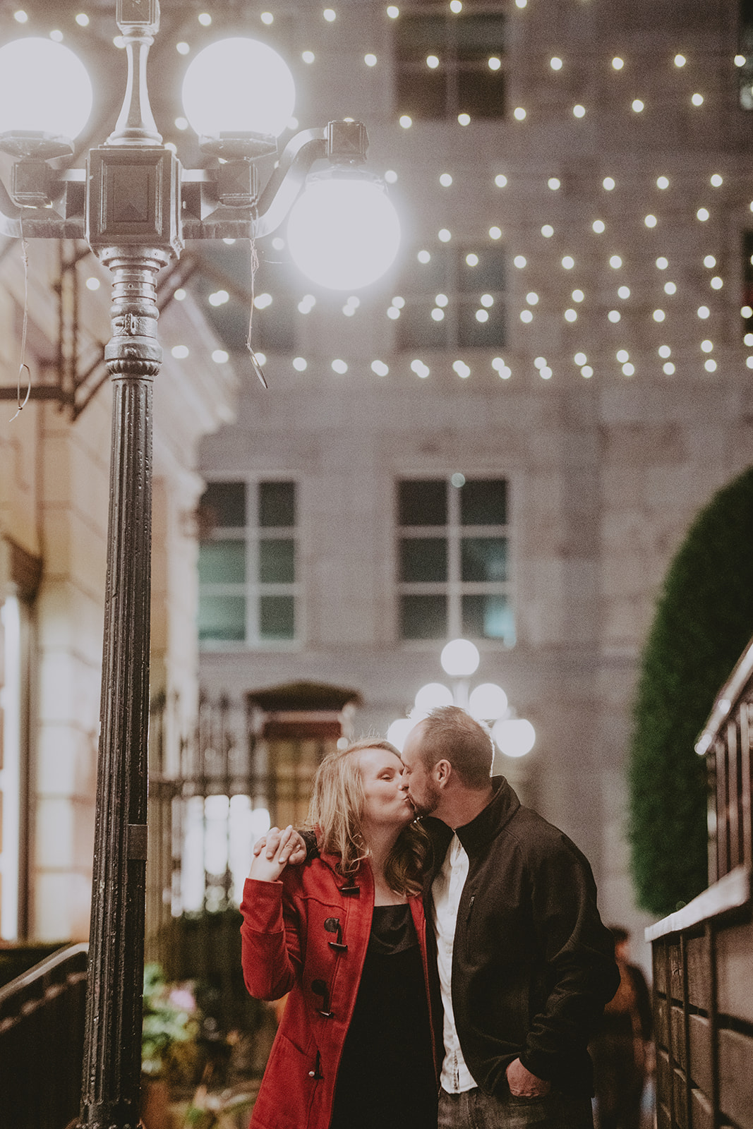 Victoria BC engagement. Woman in red jacket kissing couple in love under a lamp post. 